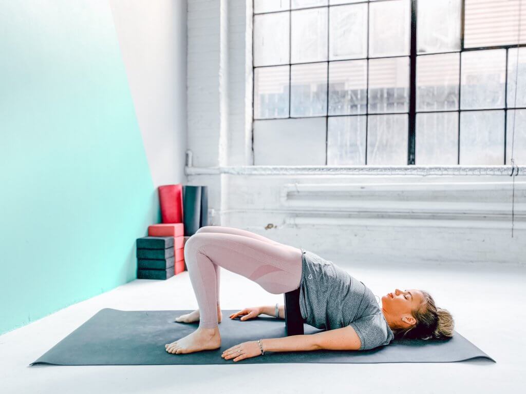 Woman in a Supported Bridge Pose with a yoga block under her sacrum.