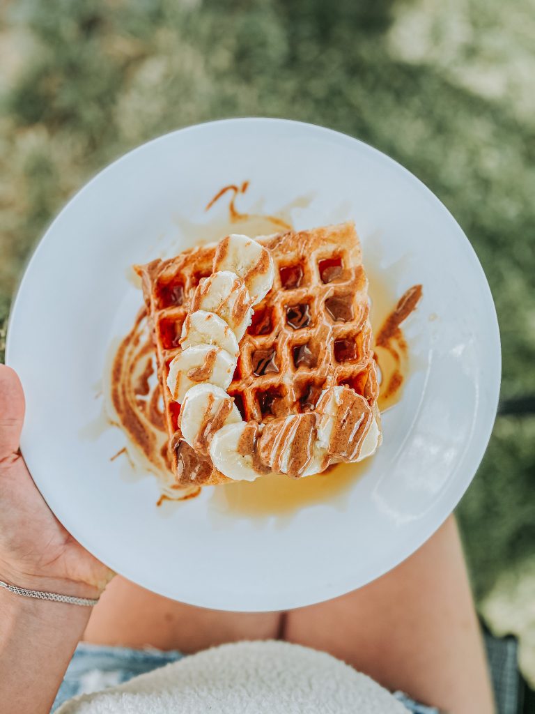 A stack of vegan waffles topped with banana slices, almond butter, and maple syrup.